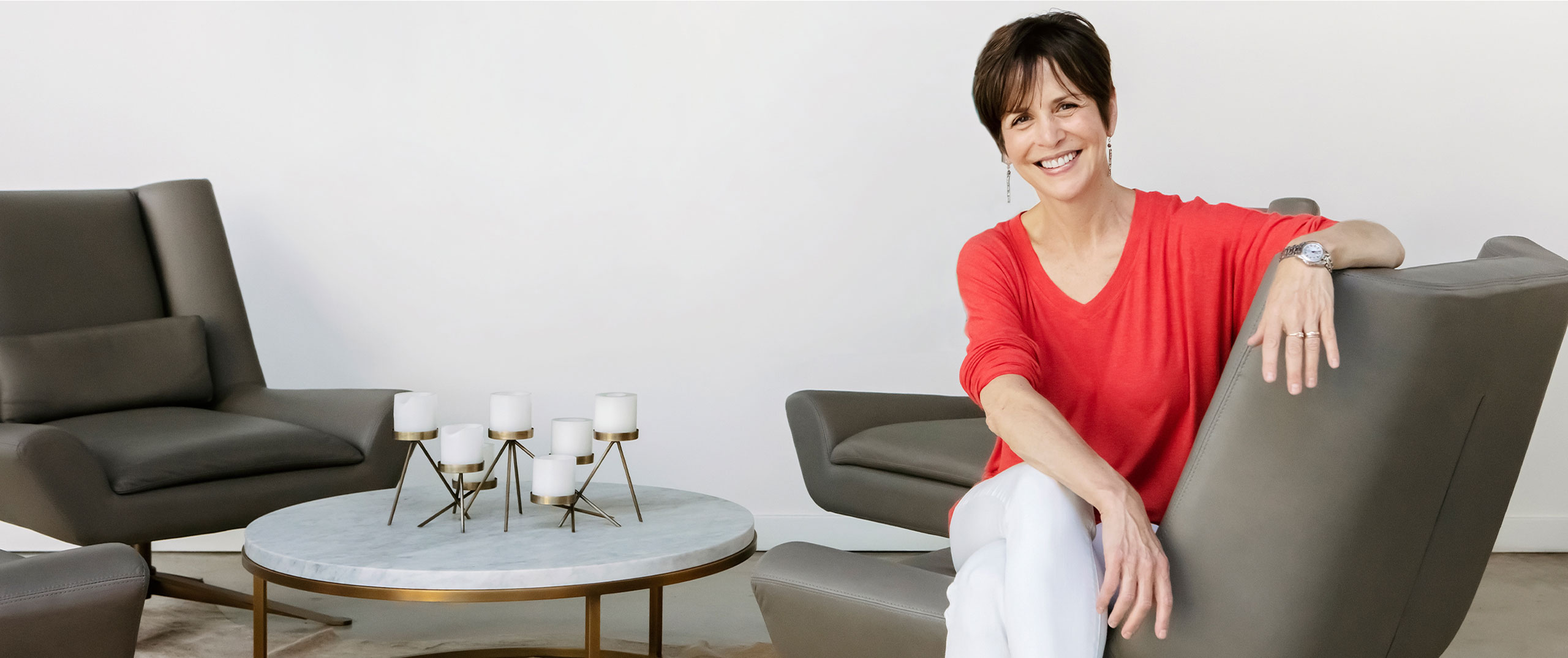 Linda Goldman sits smiling in a grey chair near a coffee table with candles on it